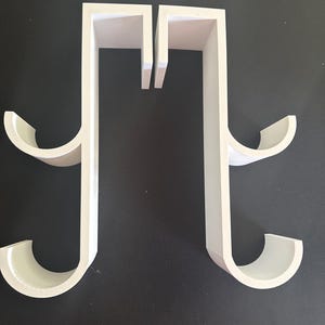 May include: Two white over-the-door hooks, each featuring a flat vertical bar with a top bracket for hanging and two curved, semi-circular hooks extending outward from the side to hold items. The structure is rigid, smooth, and matte white.