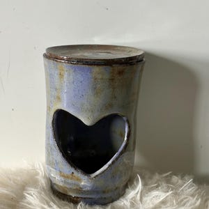 May include: A handmade ceramic jar with a lid. The jar is cylindrical with a heart-shaped cutout. It has a blue and brown glaze. The jar is sitting on a white, fluffy surface.