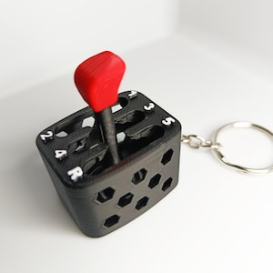 Könnte beinhalten: Ein schwarzer Schlüsselanhänger in Form eines manuellen Gangschalthebels mit rotem Knauf. Die Basis zeigt ein Schaltgitter mit den in Weiß aufgedruckten Zahlen 1, 2, 3, 4, 5 und R. Die Seiten des schwarzen Gehäuses haben ein Sechseckmuster.