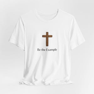 以下が含まれることがあります： 茶色の木の十字架のデザインが施された白いTシャツ。「Be the Example」の文字が十字架の下にプリントされています。Tシャツは白い生地で作られています。