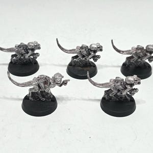 May include: Five unpainted silver-colored metal miniature figurines of reptilian creatures with long tails, each mounted on a circular black plastic base. The figures feature detailed scales, hunched postures, and distinct facial features.