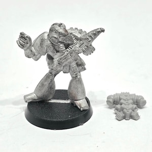 May include: A silver-colored miniature figurine of a futuristic soldier, posed with a weapon. The figure is mounted on a black base, with a separate, unattached piece nearby. The details include armor and a helmet, suggesting a science fiction or fantasy theme.
