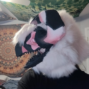 May include: A detailed, handmade dinosaur mask with a black, pink, and white color scheme. The mask features a textured, open mouth with black teeth and is surrounded by fluffy, white faux fur. The mask is designed to be worn.