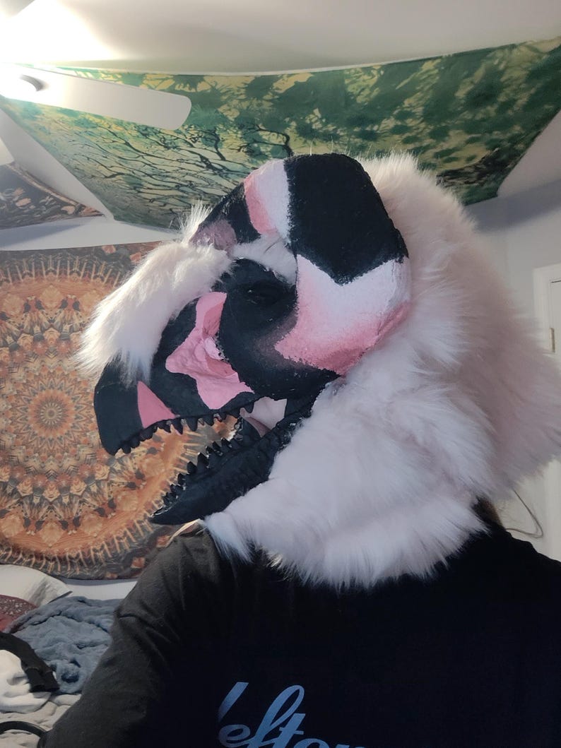 May include: A detailed, handmade dinosaur mask with a black, pink, and white color scheme. The mask features a textured, open mouth with black teeth and is surrounded by fluffy, white faux fur. The mask is designed to be worn.
