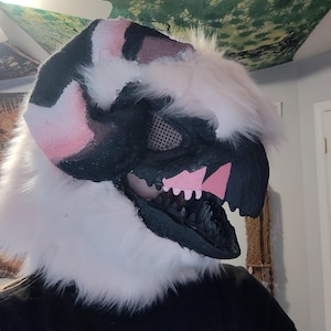 May include: A detailed close-up of a fursuit head. The head is primarily black, white, and pink, with a stylized dinosaur or dragon design. The mouth is open, revealing black teeth and pink accents. White faux fur frames the head.