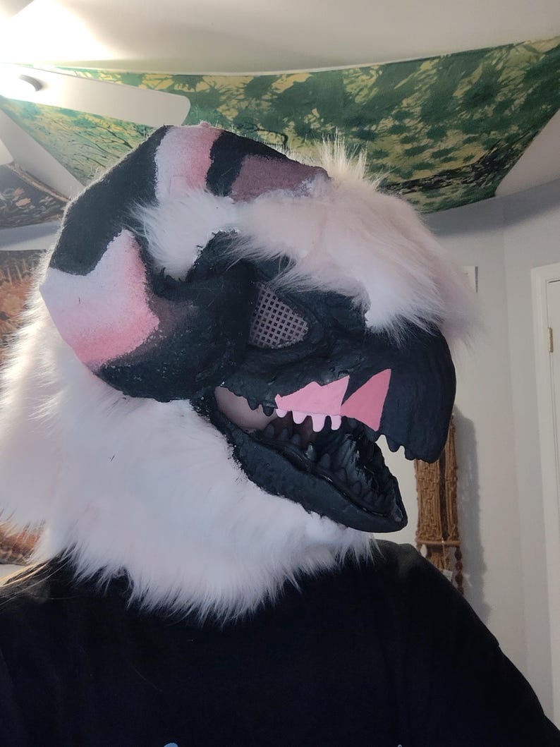 May include: A detailed close-up of a fursuit head. The head is primarily black, white, and pink, with a stylized dinosaur or dragon design. The mouth is open, revealing black teeth and pink accents. White faux fur frames the head.