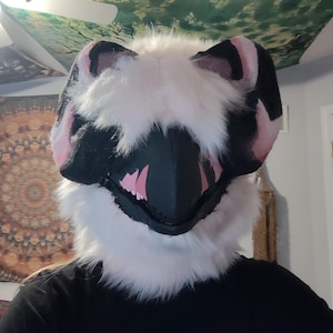 May include: A furry, stylized animal mask with a black snout and pink inner mouth. The mask has black and pink ears and is surrounded by white fur. The mask is worn by a person wearing a black shirt.