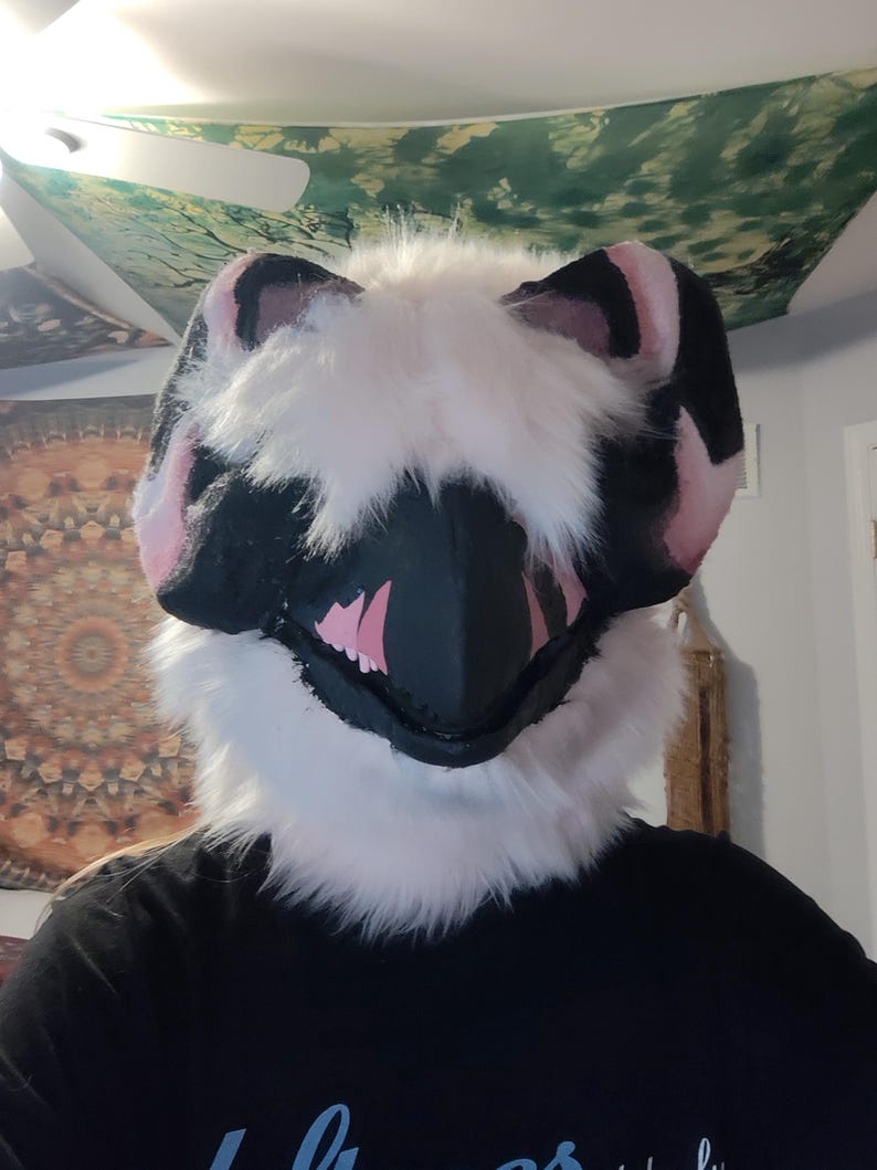 May include: A furry, stylized animal mask with a black snout and pink inner mouth. The mask has black and pink ears and is surrounded by white fur. The mask is worn by a person wearing a black shirt.