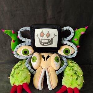 May include: A plush toy creature with a black square head featuring a pixelated face, large green eyes, and multiple appendages. The plush has a light yellow body, green and red accents, and a variety of textures.