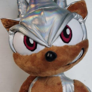 May include: A plush toy character with a silver head covering and accents, brown fur, and red-rimmed eyes. The character has a determined expression and a black nose. The toy is a soft, cuddly item.