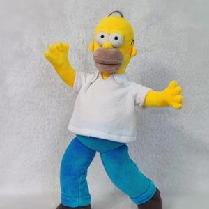 May include: A plush Homer Simpson doll with a yellow head, brown mouth, and black eyes. He wears a white shirt, blue trousers, and brown shoes. The doll has outstretched yellow arms and is posed as if walking.