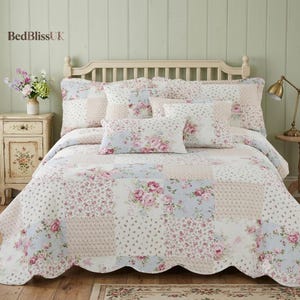 May include: A patchwork quilt and matching pillow shams featuring a floral pattern with pink roses on white, light blue, and beige backgrounds. The bedding set has a scalloped edge and is displayed on a bed with a light-coloured wooden headboard.