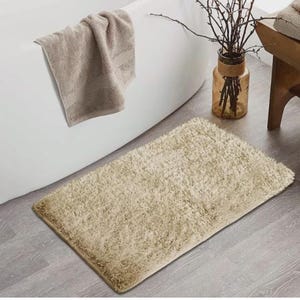 May include: A beige rectangular bath mat with a shaggy texture on a grey wooden floor. A beige towel hangs on the edge of a white bathtub. A glass vase with branches is in the background.