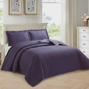 May include: A dark purple bedspread set with two matching pillowcases. The bedding has a textured, leaf-like design. The bed is made up with a white headboard, a white bedside table, and a white chest of drawers.