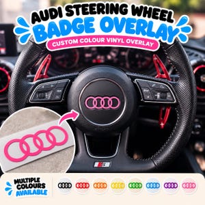 May include: A black Audi steering wheel featuring a pink vinyl badge overlay on the centre emblem. The steering wheel includes perforated leather grips, red paddle shifters, and integrated control buttons. A separate pink vinyl decal is shown.