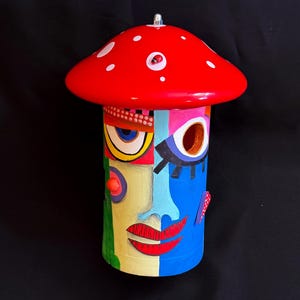 May include: A whimsical, hand-painted mushroom-shaped decorative object. The red cap features white polka dots and a small ladybug. The cylindrical body is painted with a colorful, abstract face design, including eyes, lips, and geometric patterns. The base is a mix of yellow, green, and blue.