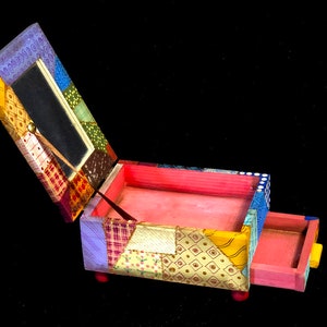 May include: A patchwork jewellery box with a hinged lid and a small drawer. The box is made of various patterned fabrics in shades of yellow, blue, red, and green. The interior is painted pink, and the drawer has a yellow knob. The box sits on small red feet.