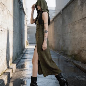 Pode incluir: Um vestido verde oliva com capuz e fenda alta, combinado com botas pretas de salto. O vestido sem mangas tem cintura com cord&atilde;o. A modelo caminha numa rua estreita com pavimento molhado.