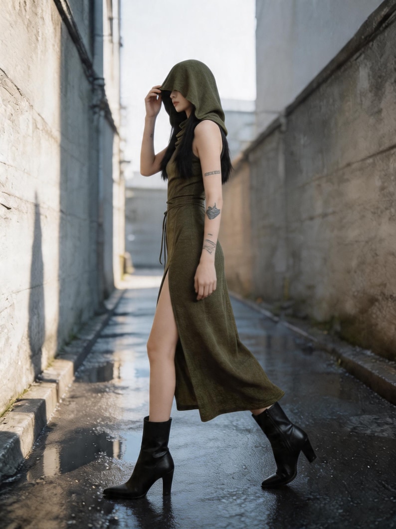 Pode incluir: Um vestido verde oliva com capuz e fenda alta, combinado com botas pretas de salto. O vestido sem mangas tem cintura com cord&atilde;o. A modelo caminha numa rua estreita com pavimento molhado.