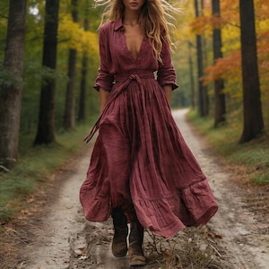 May include: A long, burgundy-colored dress with a button-down top and a flowing skirt. The dress has a waist tie and long sleeves. The person is wearing brown boots and walking on a dirt path in a forest.