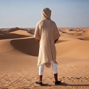 Könnte beinhalten: Eine beige Tunika mit Kapuze, kurzen Ärmeln und lockerer Passform, kombiniert mit einer weißen, knöchellangen Hose und dunklen Slipper-Schuhen. Das Kleidungsstück besteht aus einem glatten, leichten Stoff, der bis zur Mitte der Oberschenkel reicht.