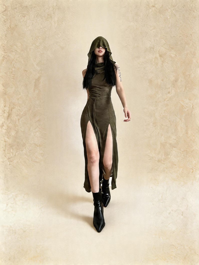 Pode incluir: Vestido verde-oliva com capuz, fendas altas e cintura com cord&atilde;o. O vestido &eacute; combinado com botins pretos. A modelo caminha para a frente sobre um fundo neutro. O vestido &eacute; sem mangas.