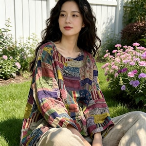 May include: A patchwork long-sleeve top in a variety of colors including red, blue, green, and yellow. The top has a round neckline and is paired with light-colored pants. The person is sitting on the grass in front of a white picket fence.