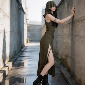 Pode incluir: Um vestido verde-oliva com capuz, fenda alta e cinto a condizer. O vestido &eacute; usado com botas pretas de salto alto. A pessoa est&aacute; num beco com pavimento molhado e paredes de bet&atilde;o.