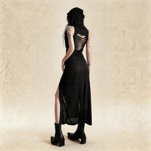 Pode incluir: Vestido preto sem mangas com capuz e fenda alta. A parte de tr&aacute;s do vestido apresenta um design recortado. A modelo est&aacute; usando botas pretas. O vestido &eacute; feito de um tecido leve.