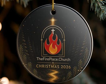 May include: A round black ceramic ornament featuring a central graphic of a red and orange flame inside an arched frame topped with a cross. The text reads TheFirePlace.Church and CHRISTMAS 2026 in gold lettering, accented by subtle gold pine tree silhouettes.