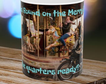 May include: A white ceramic mug featuring a photographic print of a carousel with a person riding a horse and a motorcycle parked nearby. The text "Round and Round on the Merry Go" and "are the quarters ready?" is printed in light blue letters.