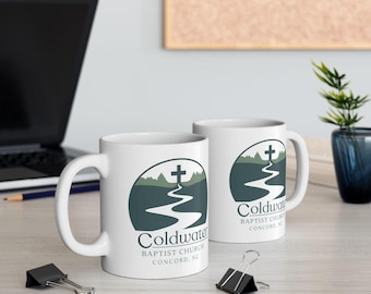 May include: Two white ceramic mugs with the text "Coldwater Baptist Church Concord, NC" printed in dark teal. The design features a cross, a winding path, and a landscape. The mugs are on a light-colored surface.