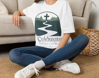 May include: White t-shirt with a graphic featuring a cross, a winding river, and the text "Coldwater Baptist Church, Concord, NC." The shirt is worn with blue jeans and white sneakers. The person is sitting on a woven rug.