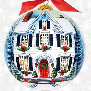 May include: A round Christmas ornament featuring a hand-painted winter scene. The ornament depicts a white house with black shutters, a red door, and a snow-covered roof. The house is decorated with wreaths and red bows. A red ribbon and gold cap are at the top.