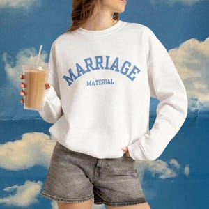 May include: A white long-sleeve crewneck sweatshirt featuring the text "MARRIAGE MATERIAL" printed in a light blue collegiate-style font across the chest. The garment is paired with gray denim shorts and is held with a clear plastic cup.