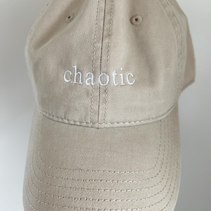 May include: A beige baseball cap featuring the word chaotic embroidered in white thread across the front panel. The cap has a structured crown, a curved brim with visible stitching, and a button at the top center of the dome.