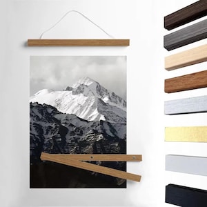 Puede incluir: Una impresión enmarcada de un paisaje de montaña cubierto de nieve, sujeta por un colgador magnético de póster de madera. El colgador es de madera de color claro y tiene un cordón blanco para colgar. Se muestran colgadores magnéticos de póster de madera adicionales en varios colores a la derecha.