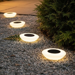 May include: Four solar-powered pathway lights illuminate a gravel path. Each light has a white, oval-shaped base with a dark, circular centre. The lights are arranged in a line, providing a soft glow.