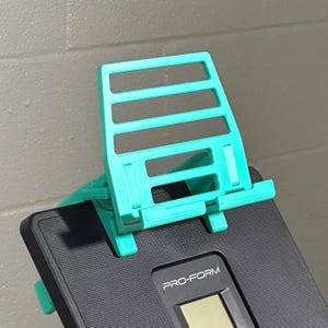 May include: A bright teal plastic tablet or phone stand with a ladder-style back support and a bottom ledge, mounted onto the black console of a Pro-Form fitness machine. The stand features a grid-like design with horizontal slats.