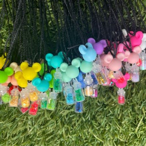 Puede incluir: Una colección de collares coloridos con colgantes en forma de Mickey Mouse en varios colores, cada uno emparejado con una pequeña botella transparente llena de purpurina. Los collares tienen cordones negros y están sobre un fondo verde.