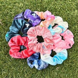 Pode incluir: Uma coleção de elásticos de cabelo de veludo em várias cores, incluindo roxo, azul-petróleo, vermelho, rosa, laranja e azul-claro. Cada elástico apresenta pequenos adornos brilhantes em forma de orelhas de rato na superfície do tecido.