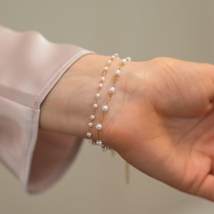May include: A delicate gold chain bracelet featuring evenly spaced small white pearls. The design consists of two thin strands of gold metal links, each adorned with round white pearls, creating a layered appearance on the wrist.