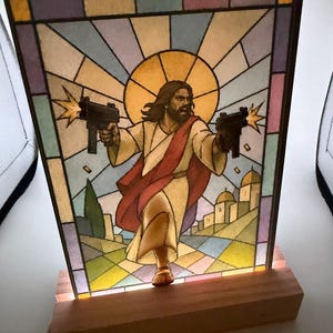May include: A rectangular light-up panel featuring a stained-glass style illustration of a figure with long hair and a red robe holding two black submachine guns with muzzle flashes. The panel is set in a rectangular wooden base with internal lighting.