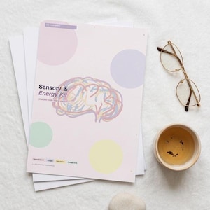 May include: A stack of white papers and a pink booklet titled "Sensory & Energy Kit" with a brain illustration and colorful circles. Gold-rimmed glasses, a small teacup, and a smooth stone are also present.