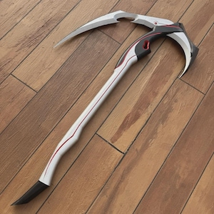 Könnte beinhalten: Zweiteilige Sense mit weißem Griff und gebogener Klinge. Der Griff hat eine rote Akzentlinie und eine schwarze Spitze. Die Klinge ist silberfarben und hat ein gebogenes Design. Die Sense liegt auf einer Holzoberfläche.