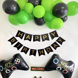 Op de afbeelding: Een feestelijke verjaardagsdecoratieset met zwarte en groene ballonnen, een banner met de tekst "HAPPY BIRTHDAY" en twee zwarte ballonnen in de vorm van een controller. Een klein bordje zegt "WELCOME".
