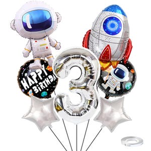 Puede incluir: Un conjunto de globos de aluminio con temática espacial que incluye un número 3 plateado grande, una figura de astronauta blanca, un cohete, dos globos redondos negros con texto Happy Birthday, dos estrellas plateadas y cinta blanca.