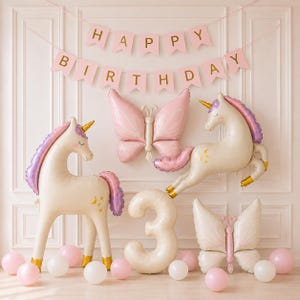 Könnte beinhalten: Ein Geburtstagsdekorationsset mit zwei großen cremefarbenen Einhorn-Folienballons mit lila Mähnen, zwei rosa Schmetterlings-Folienballons, einem großen cremefarbenen Zahlenballon drei und einem rosa Happy Birthday-Banner mit goldener Schrift.