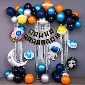 May include: A festive space-themed birthday party decoration set. Includes a variety of balloons in blue, orange, silver, and black, along with foil balloons shaped like a rocket, planets, and an astronaut. A banner reads "Happy Birthday".