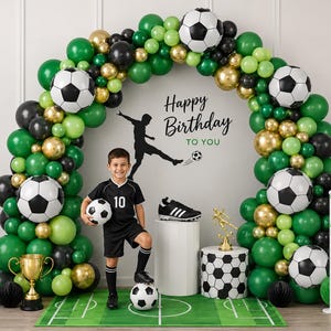 May include: A soccer-themed birthday party decoration set featuring a large balloon arch in shades of green, black, and gold, interspersed with soccer ball balloons. The display includes a Happy Birthday sign, a trophy, and a soccer field mat.